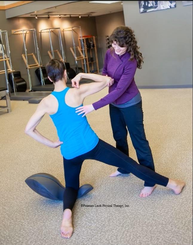 Melissa wear a purple shirt and black pants helping a young man in a blue tank top and exercise pants kneeling on an oov